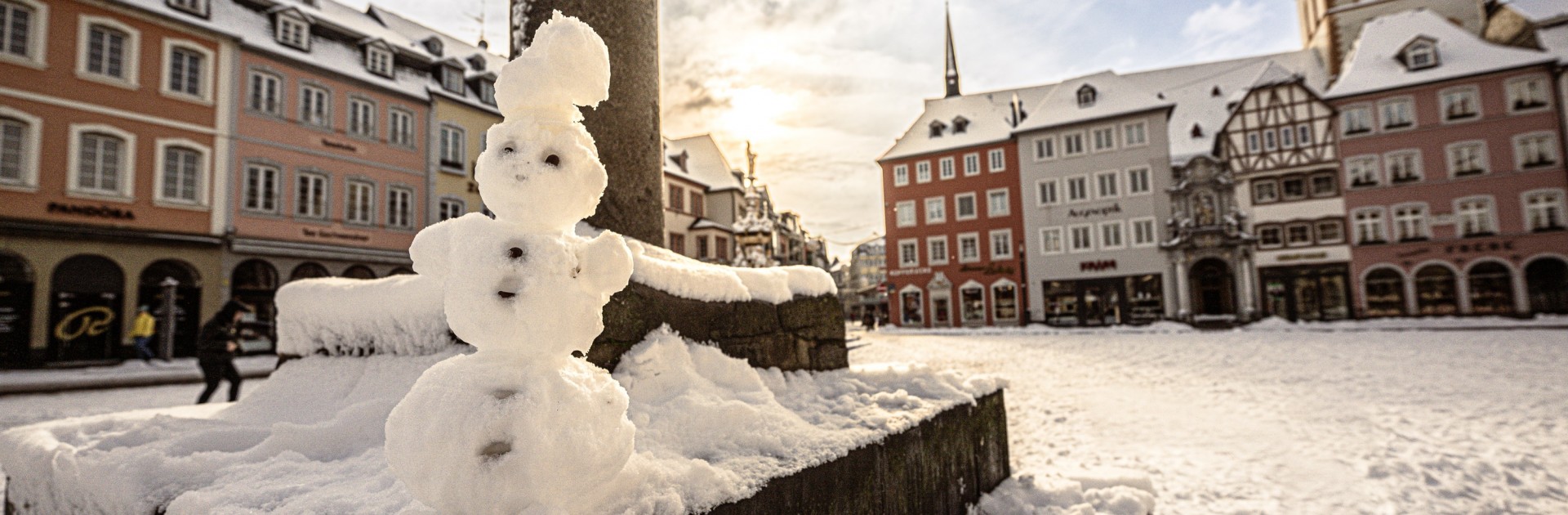 Ein selbst gebauter Schneemann steht vor dem Trierer Marktkreuz auf dem schneebedeckten Hauptmarkt. - © Trier Tourismus und Marketing/Simon Engelbert