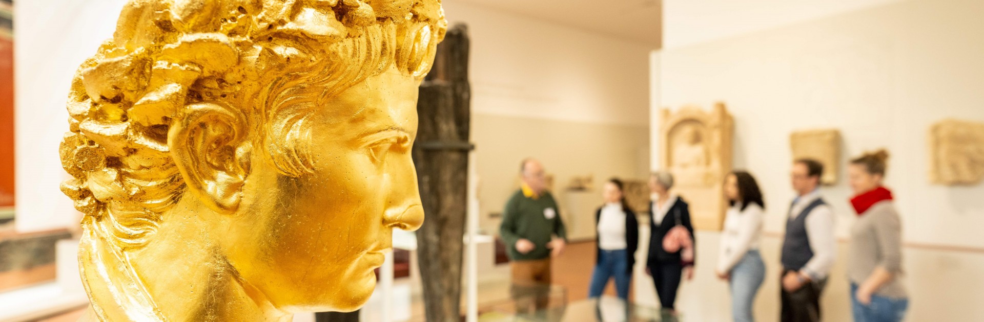 Eine Gruppe Besucher steht im Rheinischen Landesmuseum, im Vordergrund schaut ein vergoldeter Augustuskopf nach rechts. Augustus ist Stadtgründer Triers.  - © Trier Tourismus und Marketing GmbH/Dominik Ketz