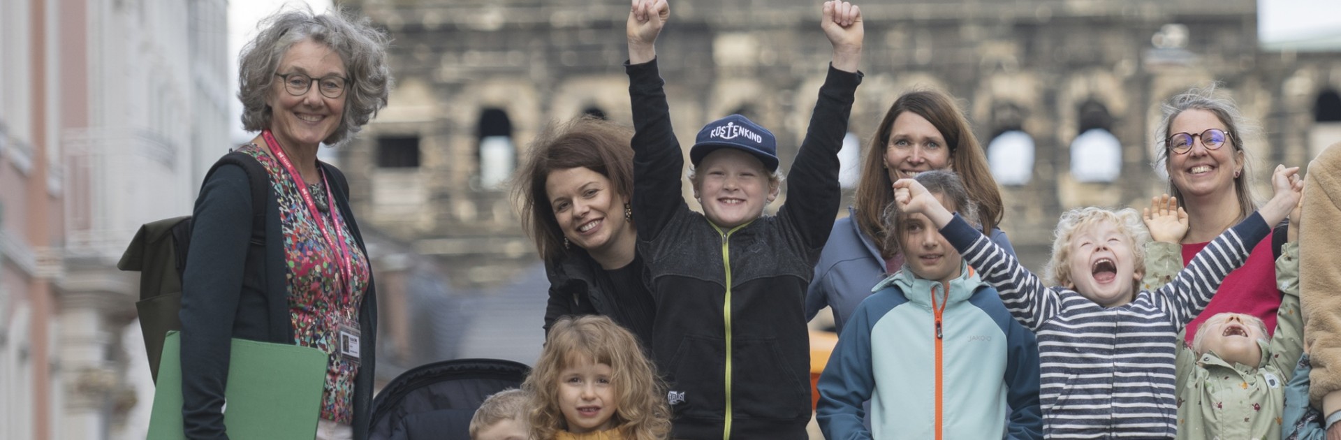Eine Familiengruppe mit kleinen und größeren Kindern steht mit einer Gästeführerin vor der Porta Nigra. Mehrere Kinder reißen jubelnd die Arme gen Himmel, die anderen Personen lächeln.  - &copy; Trier Tourismus und Marketing GmbH_Victor Beusch