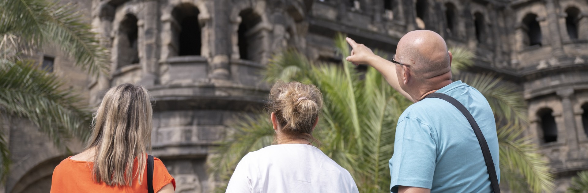 Drei Personen stehen mit dem Rücken zur Kamera vor der Porta Nigra in Trier. Ein Mann im türkisfarbenen T-Shirt zeigt nach oben auf das antike römische Stadttor; neben ihm stehen zwei Frauen in orangefarbenem und weißem T-Shirt. - &copy; Trier Tourismus und Marketing_Victor Beusch