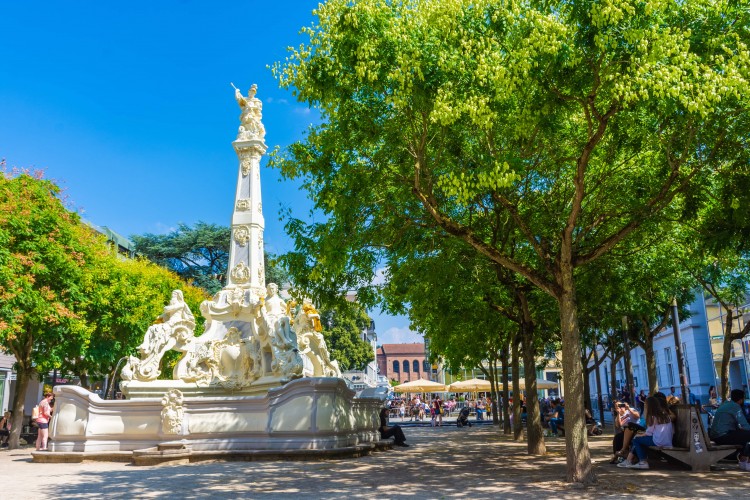 St George's Fountain on the Kornmarkt with trees, benches in the shade and the Basilica of Constantine in the background - © stock.adobe.com/Stefano Zaccaria