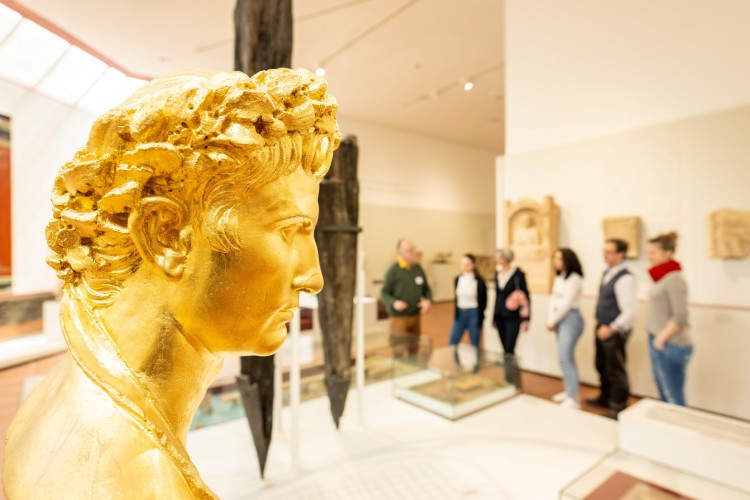 Eine Gruppe Besucher steht im Rheinischen Landesmuseum, im Vordergrund schaut ein vergoldeter Augustuskopf nach rechts. Augustus ist Stadtgründer Triers.  - © Trier Tourismus und Marketing GmbH/Dominik Ketz