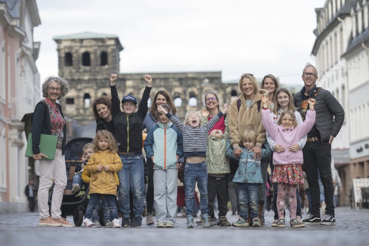 Eine Familiengruppe mit kleinen und größeren Kindern steht mit einer Gästeführerin vor der Porta Nigra. Mehrere Kinder reißen jubelnd die Arme gen Himmel, die anderen Personen lächeln.  - &copy; Trier Tourismus und Marketing_Victor Beusch