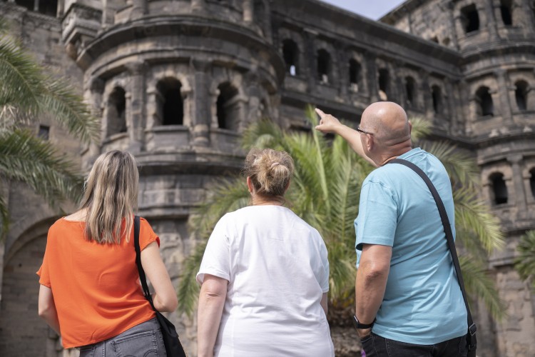 Drei Personen stehen mit dem Rücken zur Kamera vor der Porta Nigra in Trier. Ein Mann im türkisfarbenen T-Shirt zeigt nach oben auf das antike römische Stadttor; neben ihm stehen zwei Frauen in orangefarbenem und weißem T-Shirt. - &copy; Trier Tourismus und Marketing_Victor Beusch